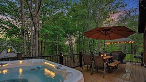 A hot tub on a wooden deck with a pergola and string lights, surrounded by trees.