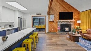 A modern kitchen with a central island, a dining area with yellow chairs, a fireplace, and a TV mounted on the wall.