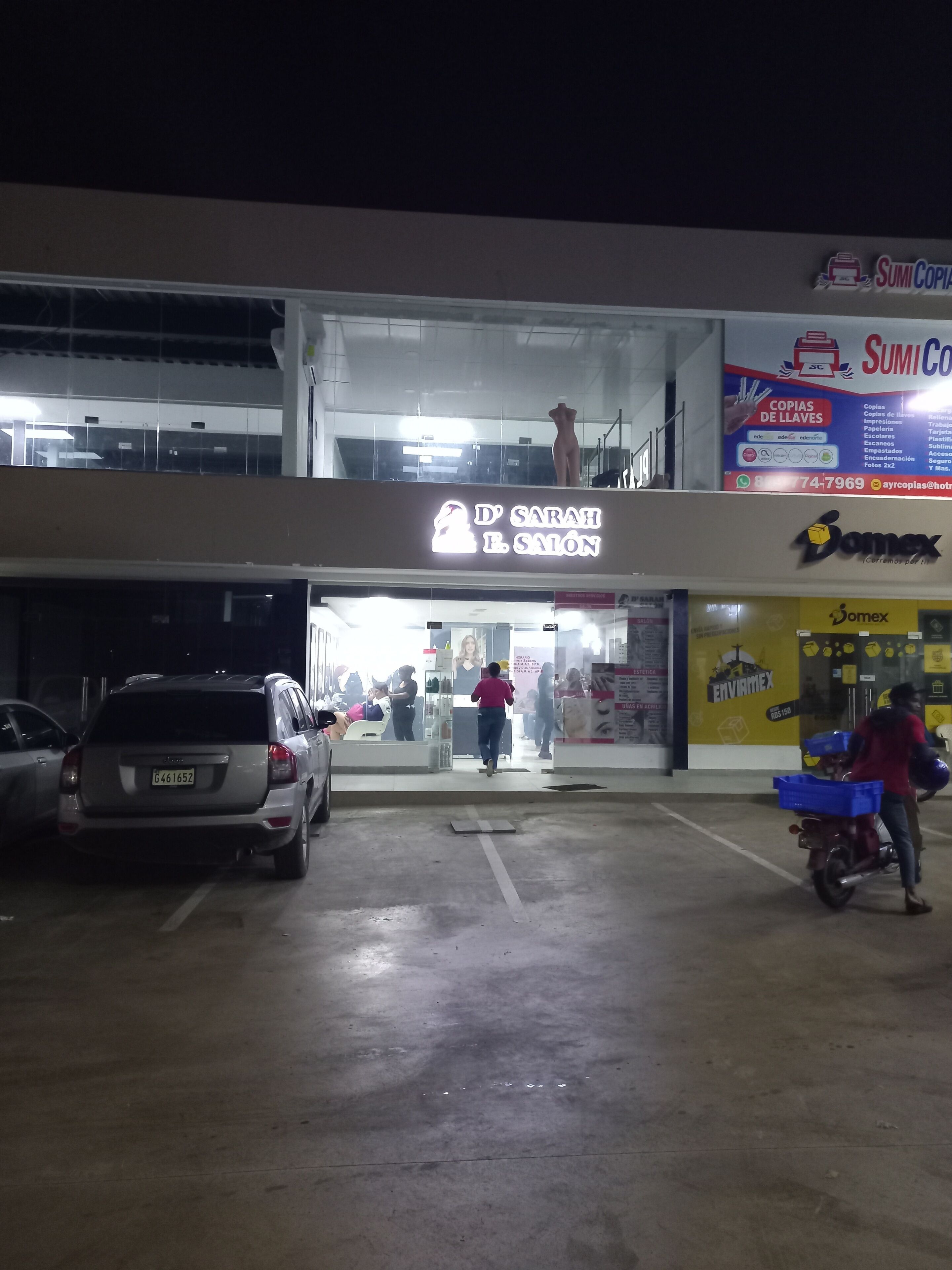 A building with a sign for 'D'Sarah B. Salon' and 'Domex' on the ground floor. There are cars parked in front and a person riding a motorcycle with a delivery box.
