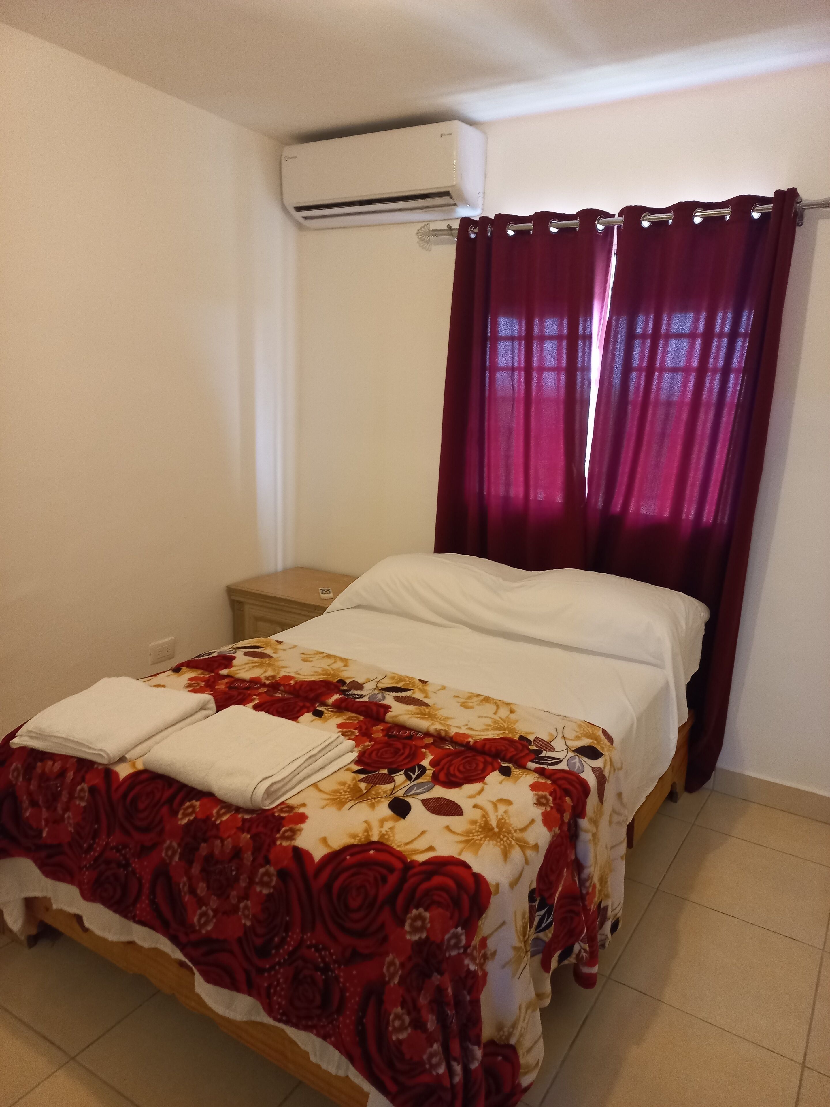 Una cama individual con una colcha estampada, un almohadón blanco y dos toallas dobladas. A la izquierda de la cama hay una mesita de noche y una ventana con cortinas rojas. Un equipo de aire acondicionado está montado en la pared sobre la cama.