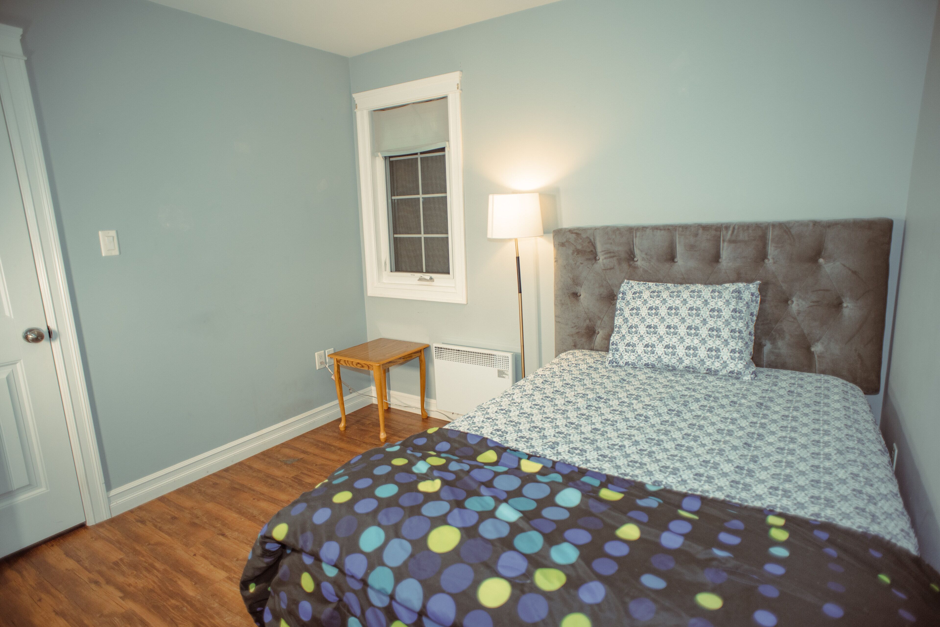 A bedroom with a bed, a nightstand, a window, and a wall-mounted lamp.