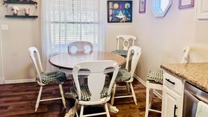 Een eetgedeelte met een ronde tafel, witte stoelen, een hanglamp en een raam met dunne gordijnen.
