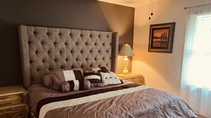 Een slaapkamer met een gestoffeerd hoofdeinde, een bedlampje, een ingelijste foto aan de muur en een raam met gordijnen.