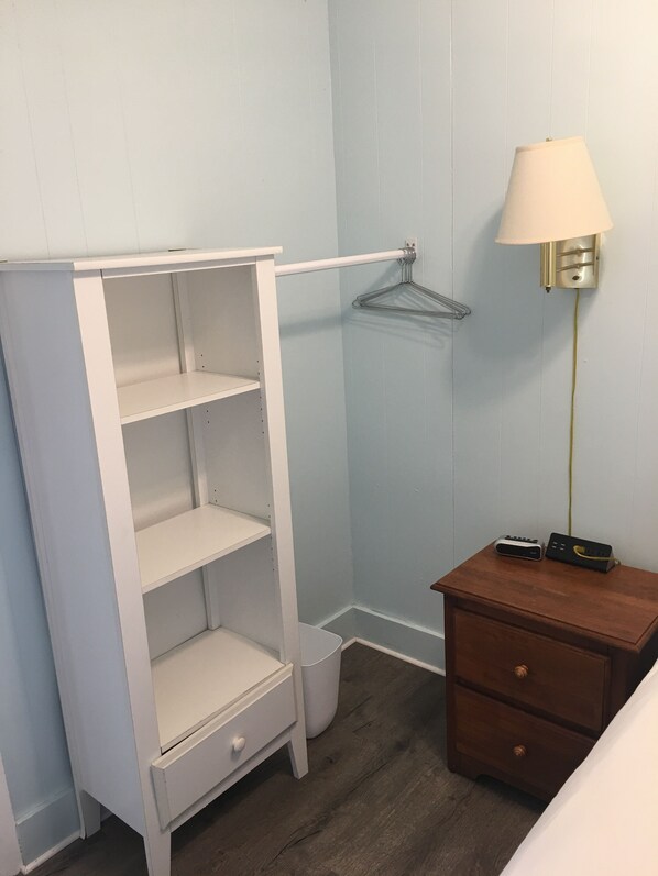 A room with a white wardrobe, a wooden nightstand, a lamp, and a hanging clothes rack.