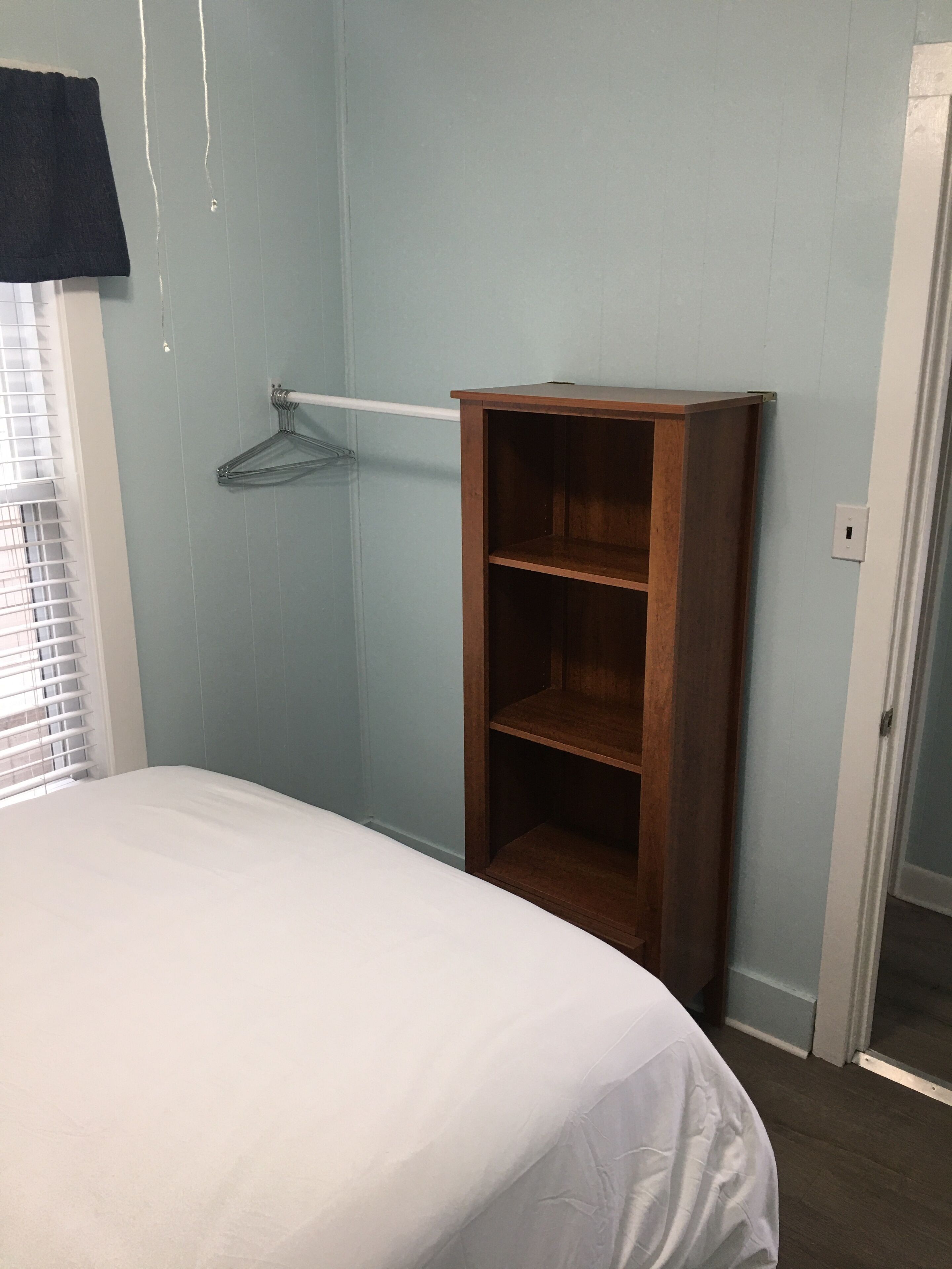 A bedroom with a bed, a wooden wardrobe, a hanging rack, and a door.