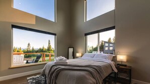 Quarto com uma cama grande, duas luminárias de cabeceira, vista para um terraço e montanhas, e uma janela com vista para um edifício.