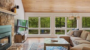Un salon spacieux avec une cheminée en pierre, de grandes fenêtres donnant sur la forêt, un canapé confortable et une table basse en verre.