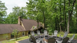 Un chalet doté d’une cheminée en pierre, entouré d’une forêt. On y trouve un foyer avec des chaises disposées tout autour.