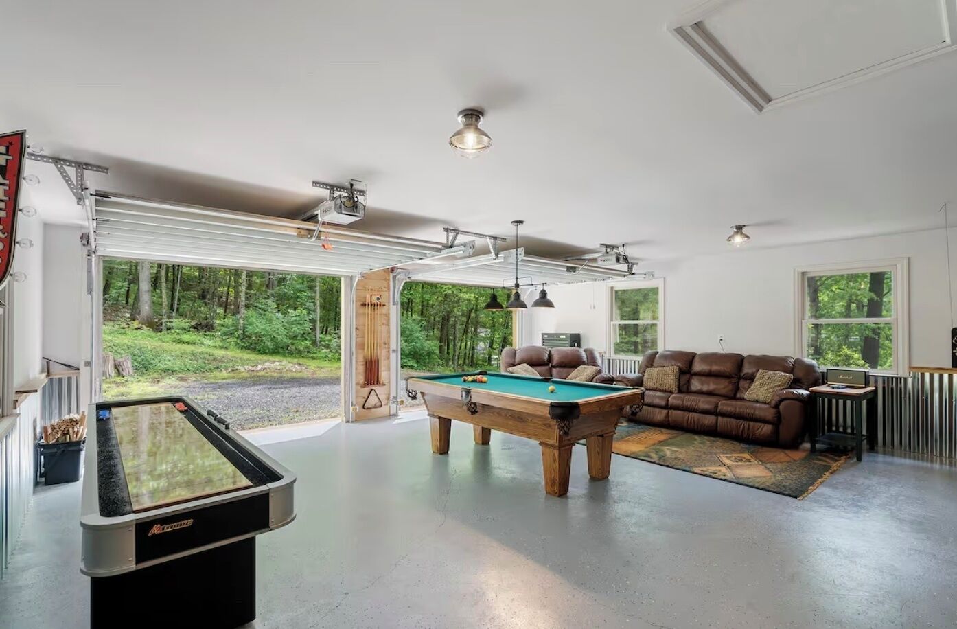 A spacious living area with a pool table, a brown leather couch, and a view of a forest through large windows.