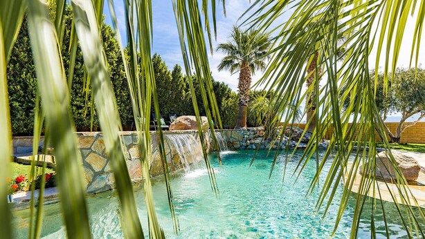 A clear view of a swimming pool with a waterfall feature, surrounded by palm trees and a stone wall. - NEW: Pool, Spa, Tiki Bar, Mini Golf, Firepit, Desert Views (Desert Hot Springs)