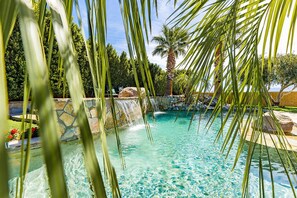 A clear view of a swimming pool with a waterfall feature, surrounded by palm trees and a stone wall. - NEW: Pool, Spa, Tiki Bar, Mini Golf, Firepit, Desert Views (Desert Hot Springs)