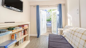 Quarto com televisão de tela plana na parede, estante com diversos livros, sofá cinza e janela com cortinas azuis.