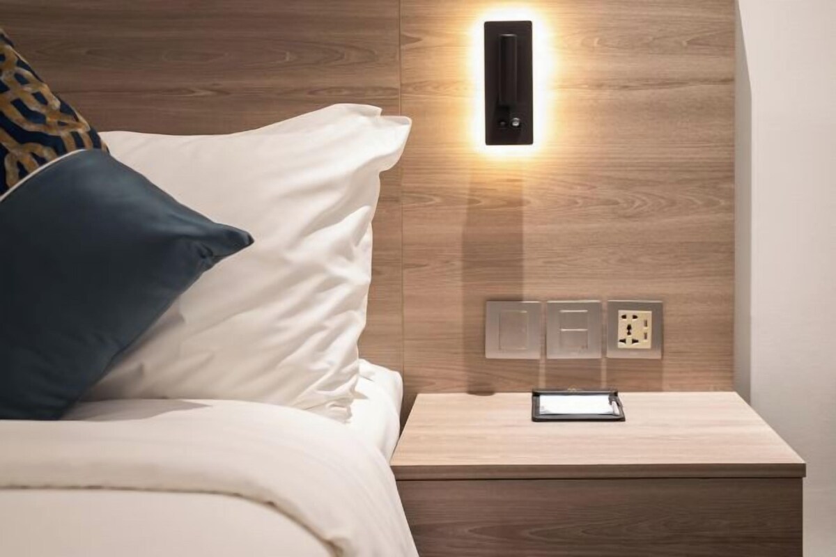 A neatly made bed with white and dark blue pillows, a bedside table with a tablet, and a wall-mounted light fixture.