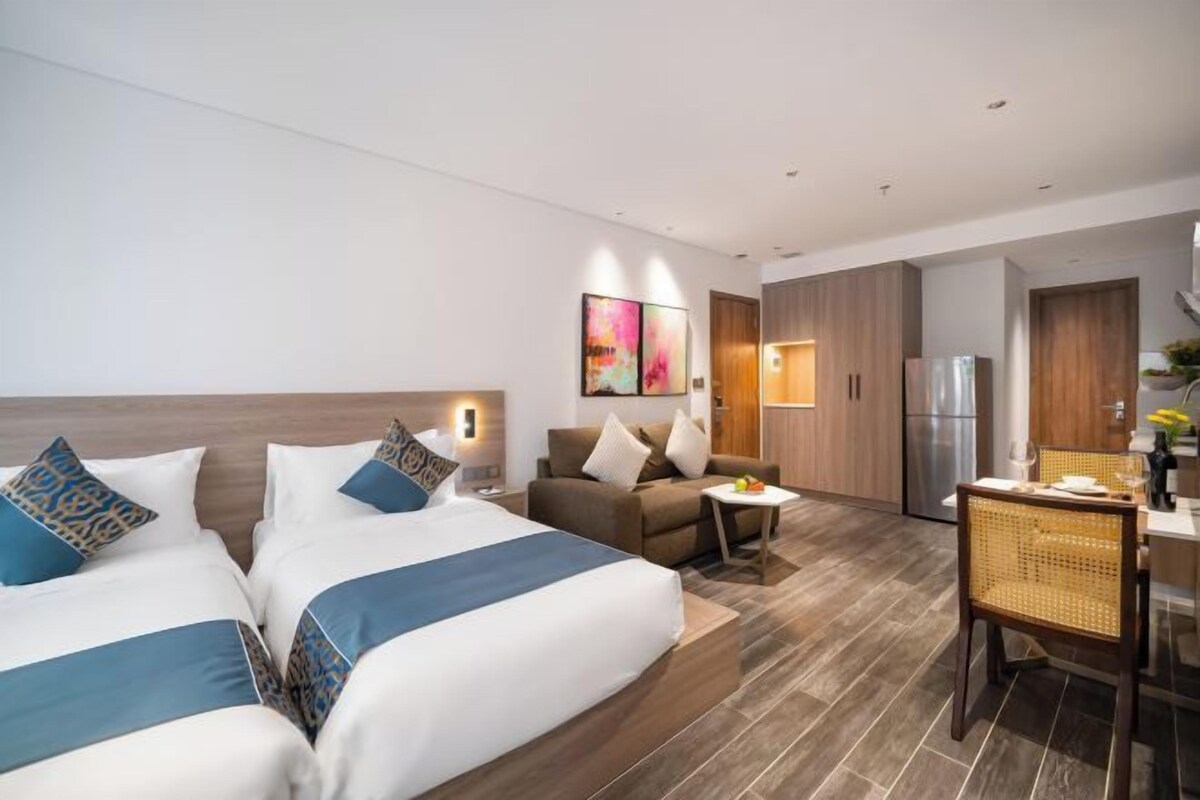 A modern hotel room with two beds, a sofa, a dining area, and a kitchen.