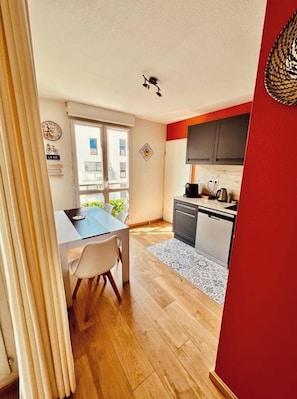 A modern kitchen with a red accent wall, a dining area with a table and chairs, and a view of the city through large windows.