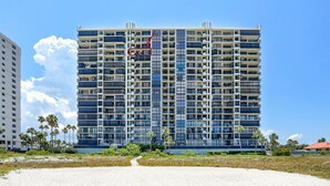 Un hôtel de grande hauteur doté de nombreux balcons, avec une plage de sable au premier plan et des palmiers.
