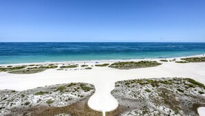 Une plage de sable blanc avec une végétation clairsemée, un ciel bleu dégagé et des eaux océaniques calmes.