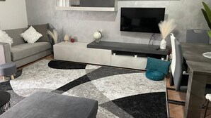 A modern living room with a grey sofa, a coffee table, a TV mounted on a wall, and a dining area with a table and chairs.