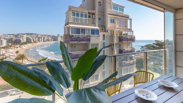 Ein Balkon mit Blick auf Strand und Meer, ein Tisch mit Tellern und üppige, grüne Pflanzen.