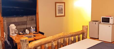Un dormitorio con estructura de cama de madera, un escritorio con cafetera, un microondas y un cuadro en la pared.