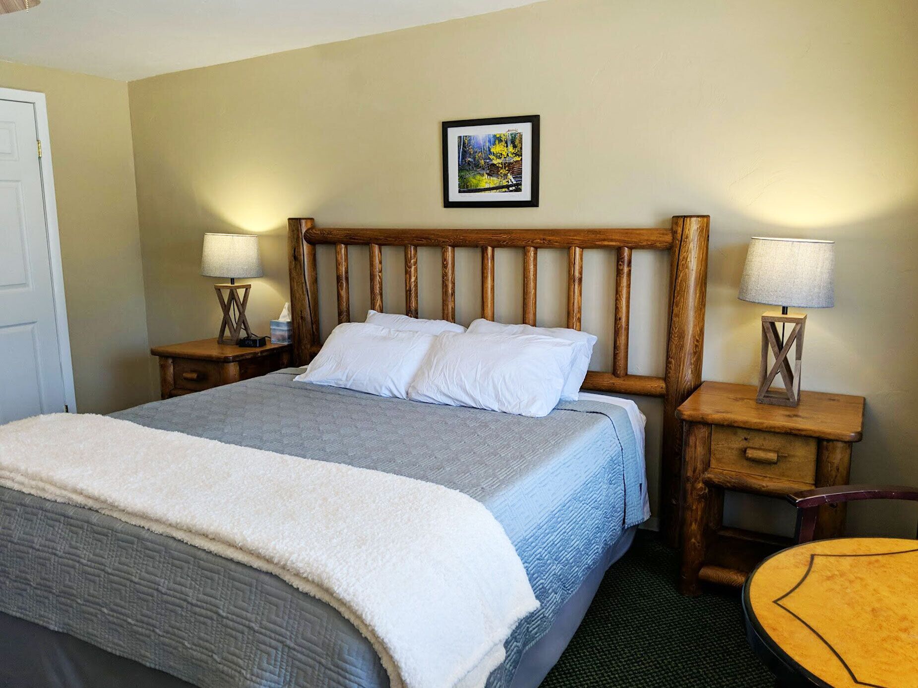 A neatly made bed with a wooden headboard, two bedside tables with lamps, a framed picture on the wall, and a small round table.