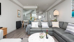 Ein modernes Wohnzimmer mit grauem Sofa, rundem Couchtisch mit Flasche und Snacks, einem Essbereich mit Tisch und Stühlen sowie einer Treppe zu einer weiteren Ebene.