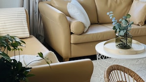 Een woonkamer met een beige bank, een witte fauteuil, een ronde salontafel en een rieten stoel. Op de salontafel staat een vaas met bloemen.