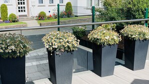 Een balkon met bloeiende planten en uitzicht op een gebouw met een witte gevel en een groene omheining.