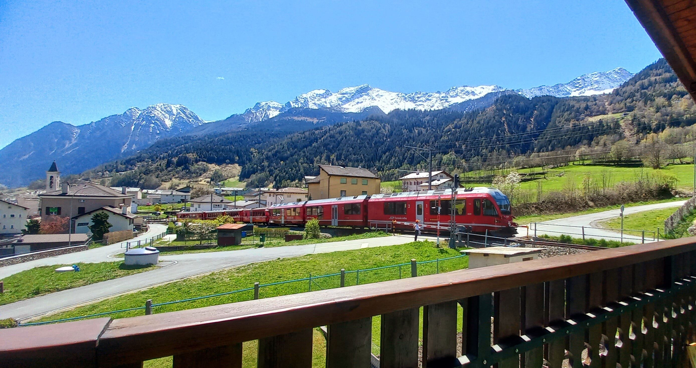Un tren rojo pasando por un pequeño pueblo con montañas al fondo.