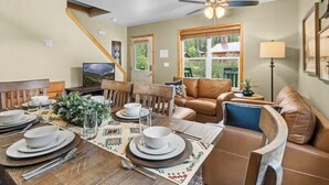 Un coin repas avec une table en bois dressée pour quatre personnes, un canapé en cuir marron et un ventilateur de plafond.