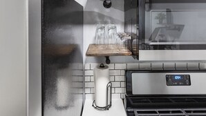 Une cuisine équipée d’un four en acier inoxydable, d’une horloge numérique affichant 10h54, et d’une étagère où sont rangés des verres et une machine à café.
