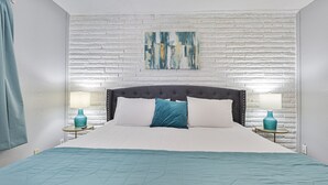 Une chambre avec un mur en briques blanches, une tête de lit rembourrée grise, une couvre-lit matelassée de couleur turquoise, deux tables de chevet avec des lampes turquoise et un tableau au-dessus du lit.