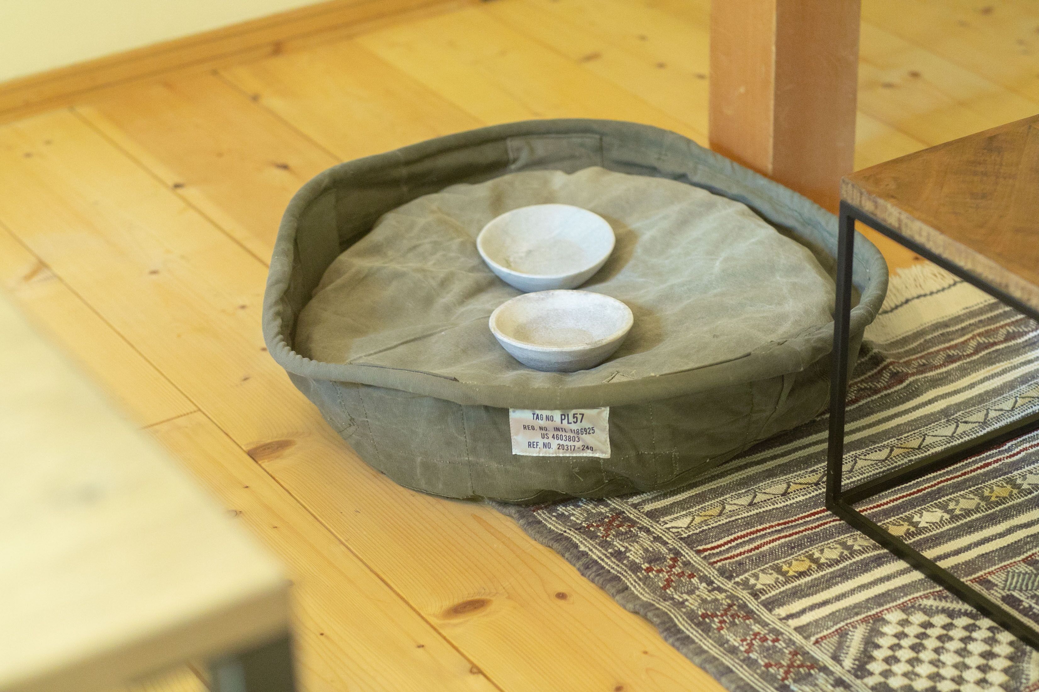 Una cama gris para mascotas con dos cuencos sobre un suelo de madera, una alfombra estampada y el pie de una mesa de metal.
