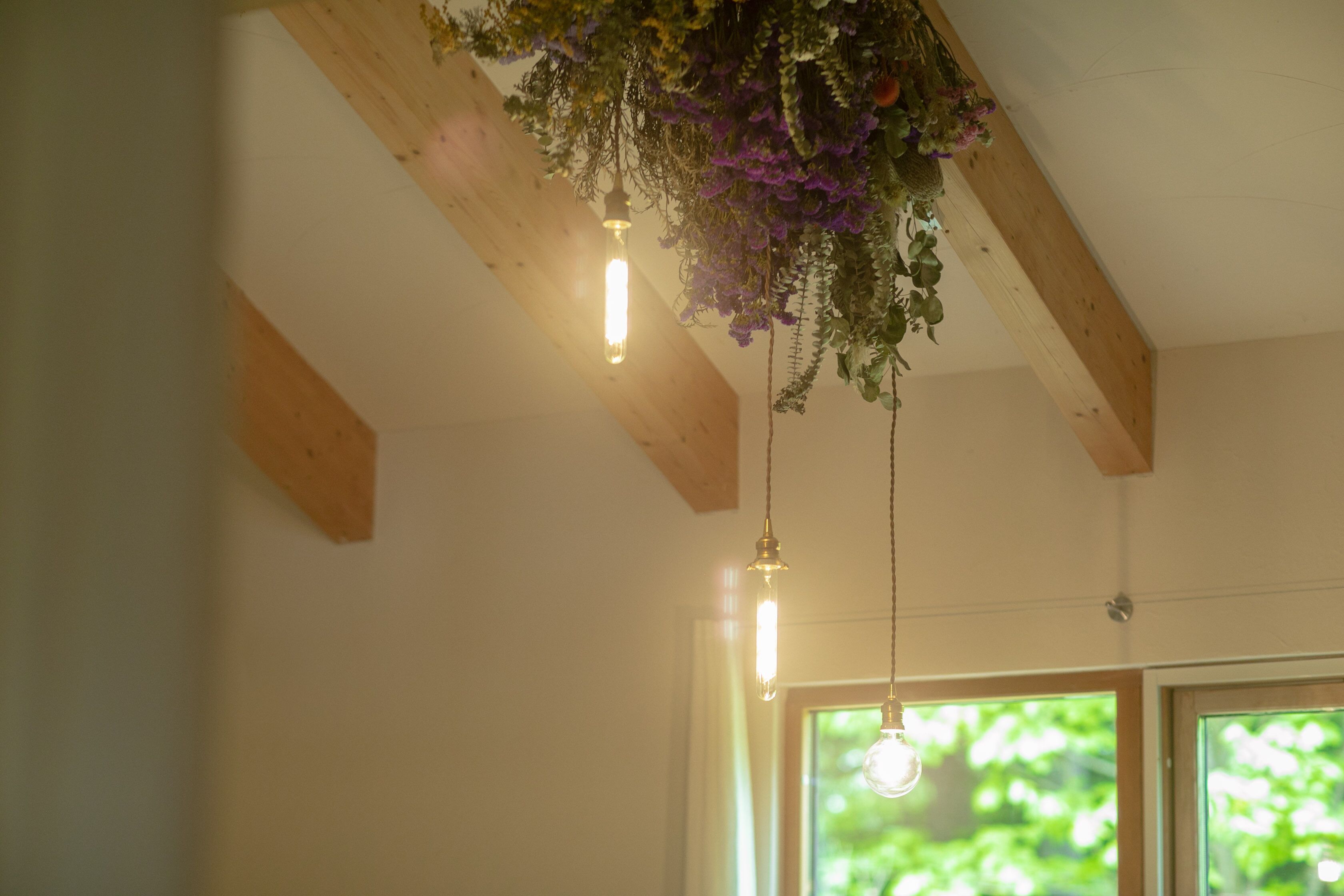 A room with exposed wooden beams, hanging plants, and a window with a view of greenery.