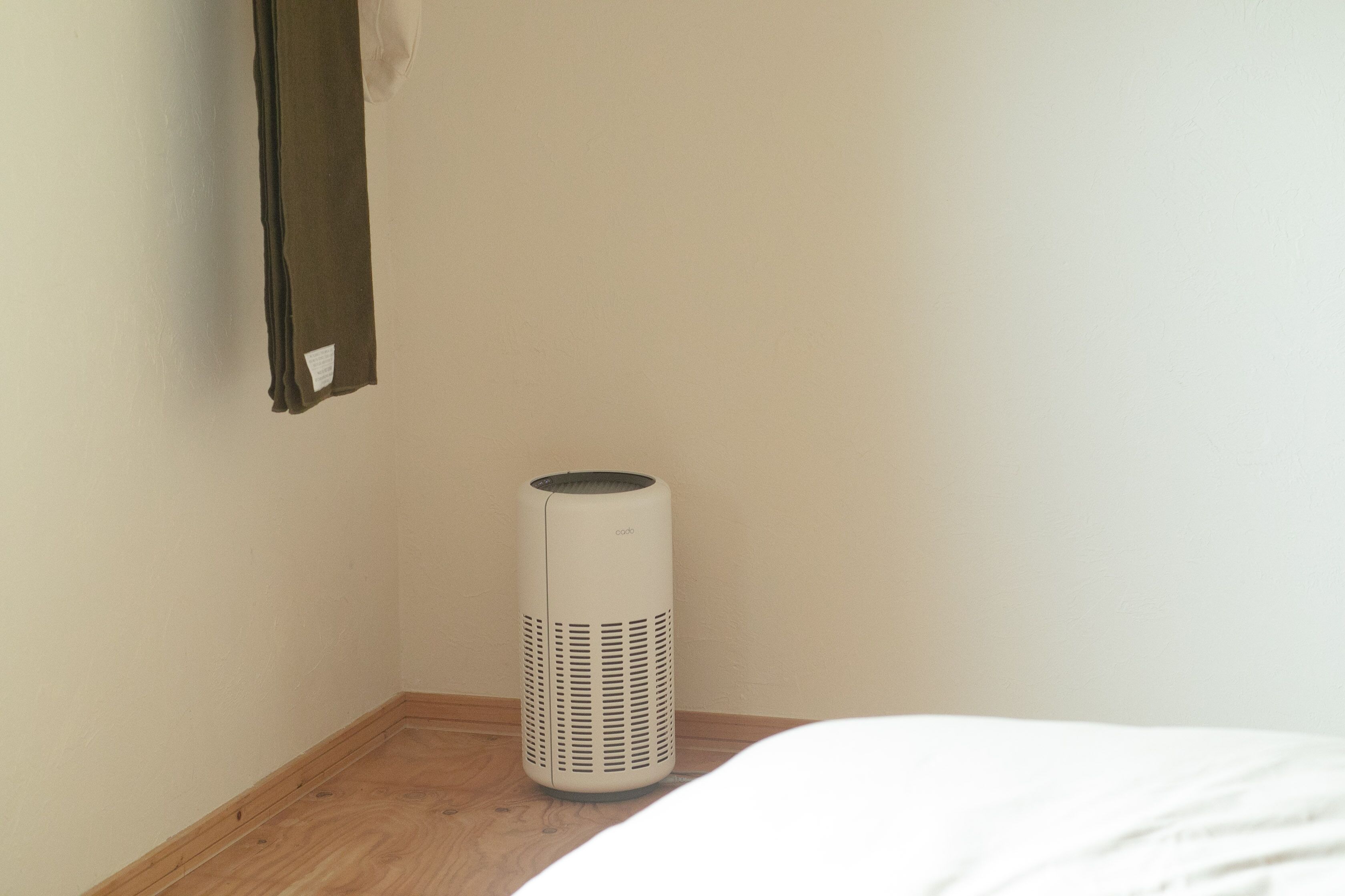 A white portable heater on a wooden floor next to a bed with a white mattress.