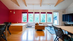 A spacious room with a high ceiling, wooden beams, a red accent wall, a large window with a view of trees, a sofa, a table, and a TV. - THE FAM (Nakafurano)