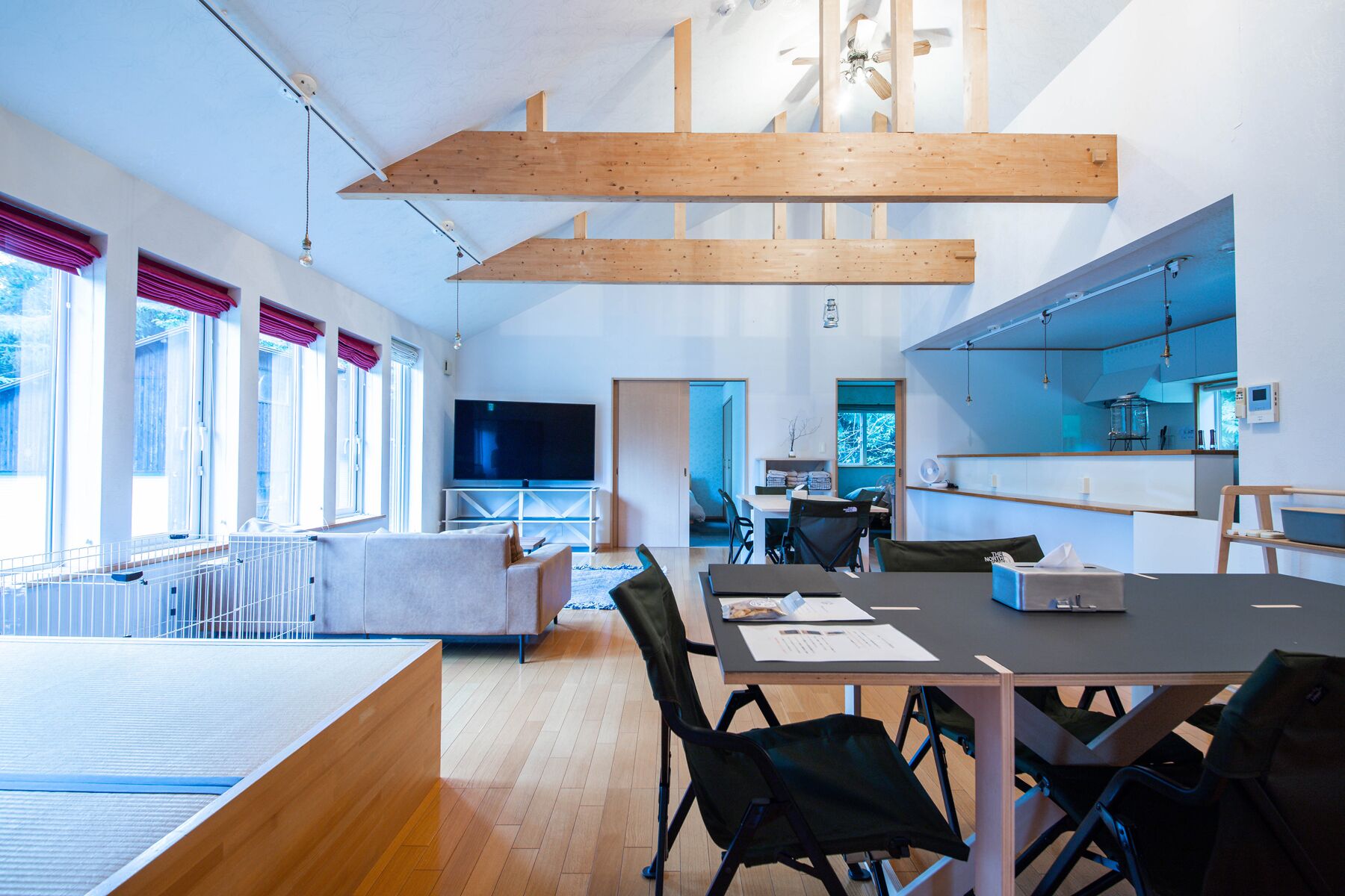 Una sala de estar moderna con un televisor grande, vigas de madera, un comedor y una cocina con iluminación azul.