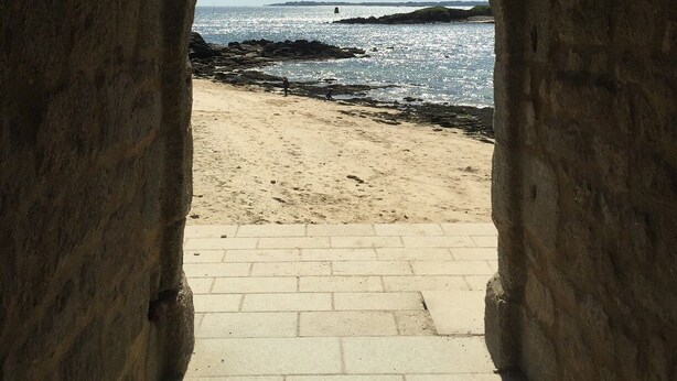 Une structure en pierre en forme d’arche mène à une plage de sable et offre une vue sur la mer.