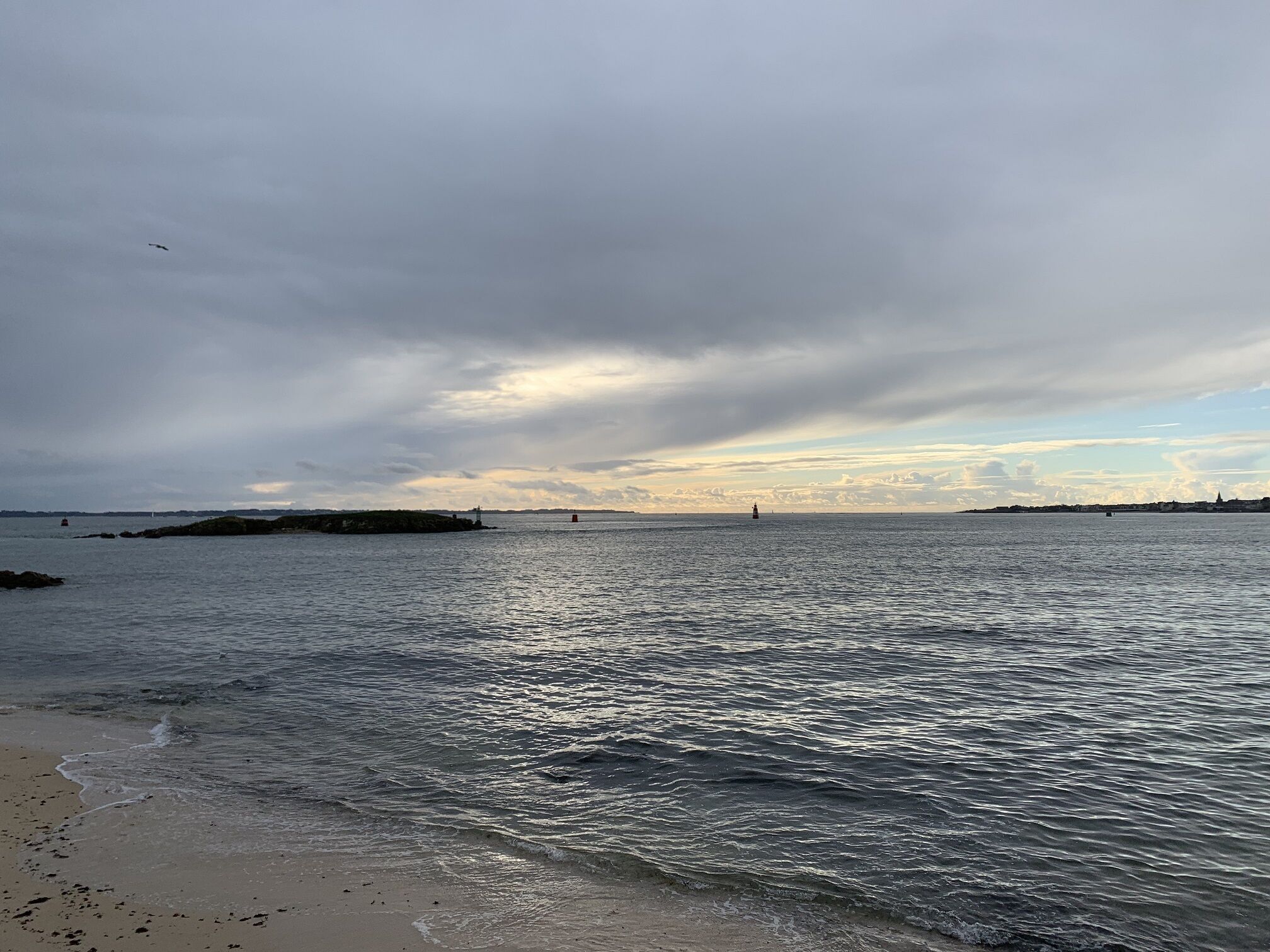 Una platja amb aigües tranquil·les, un far a la distància i un cel ennuvolat.