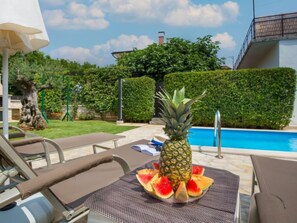 Zona junto a la piscina con sillas de descanso, una mesa con rodajas de piña y sandía, y vistas a la piscina y vegetación.
