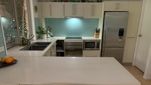 Una cocina moderna con armarios blancos, un refrigerador de acero inoxidable, un microondas y un lavamanos. Hay dos sillas blancas en la isla de la cocina.