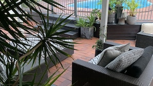 Un espacio de estar al aire libre cubierto con un sofá de mimbre y cojines, rodeado de plantas en macetas y vistas a una piscina.