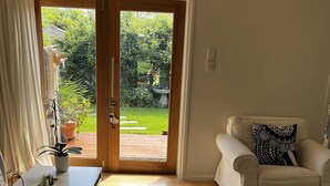 Una habitación con un sofá blanco, una pequeña mesa con vasos y una bandeja, un tapete grande tejido y un cerramiento de vidrio corredero que da a un jardín.