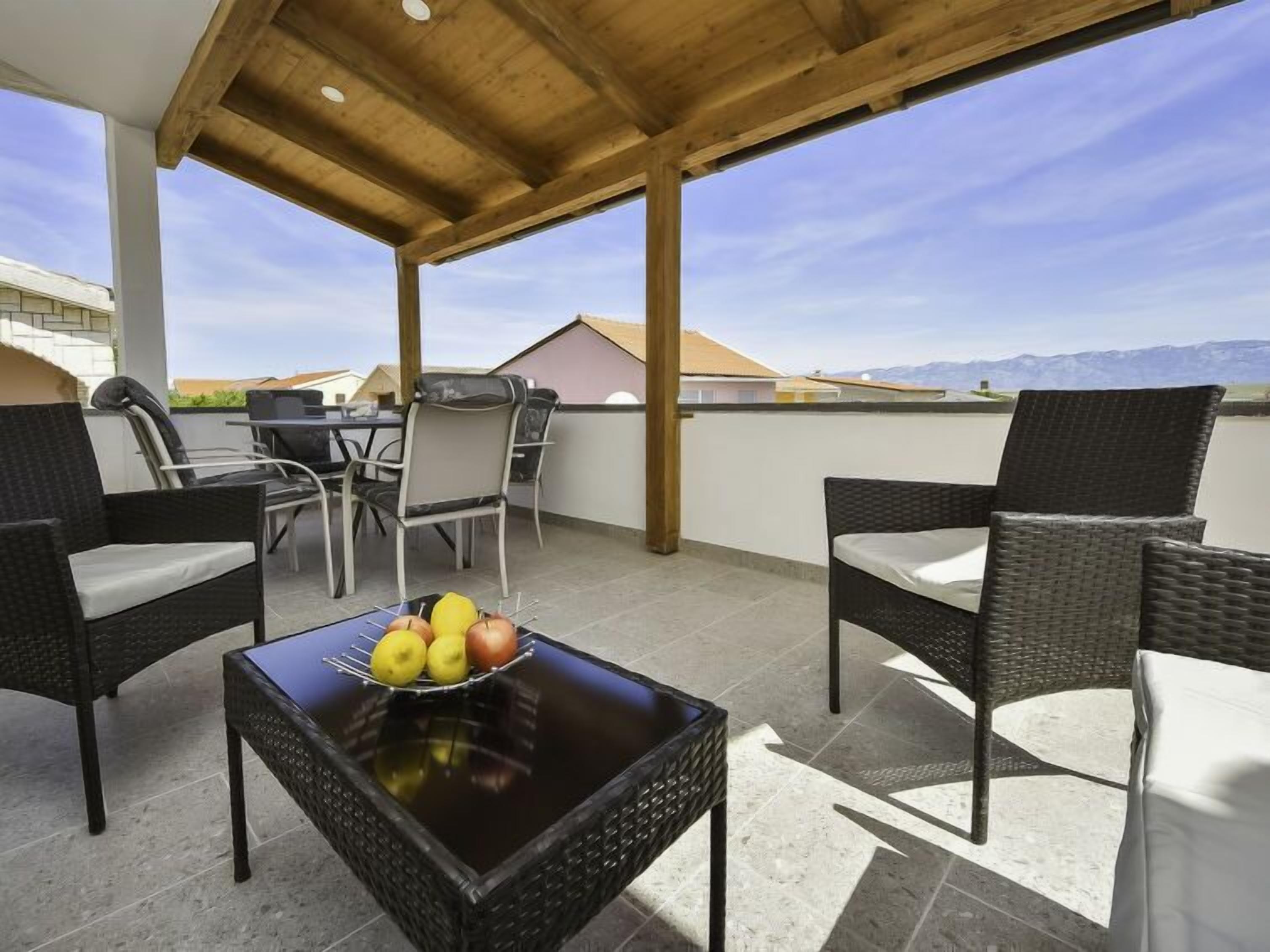 A covered outdoor seating area with wicker furniture, a table with fruit, and a view of mountains.