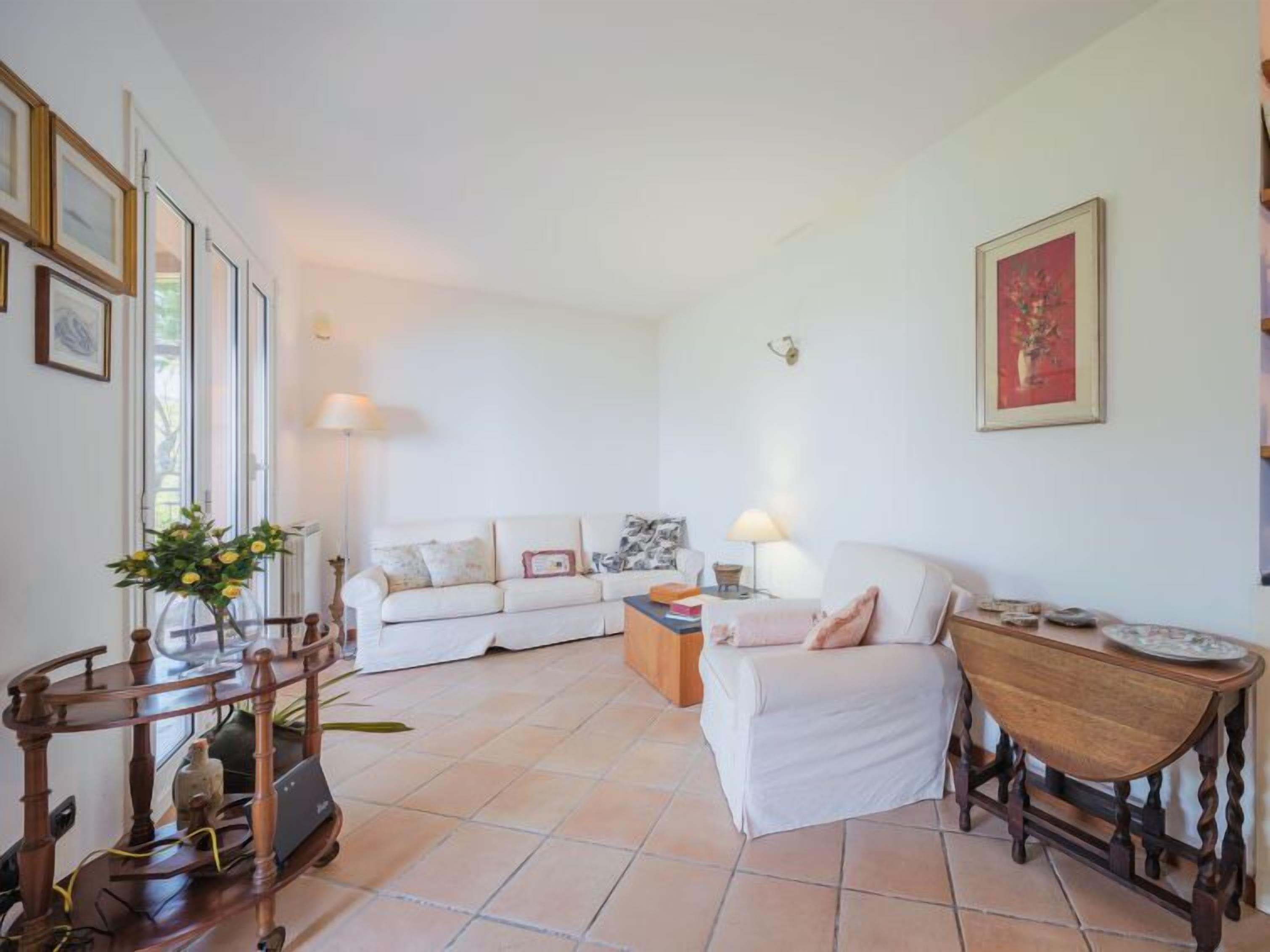 Un salón con un sofá blanco, una mesa de centro de madera y una mesita auxiliar. Hay cuadros enmarcados en la pared y un jarrón con flores sobre la mesa de centro.