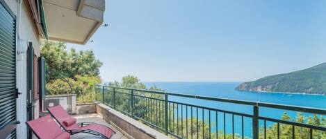 A balcony with two red lounge chairs, a tiled floor, and a view of the sea and a green hill.