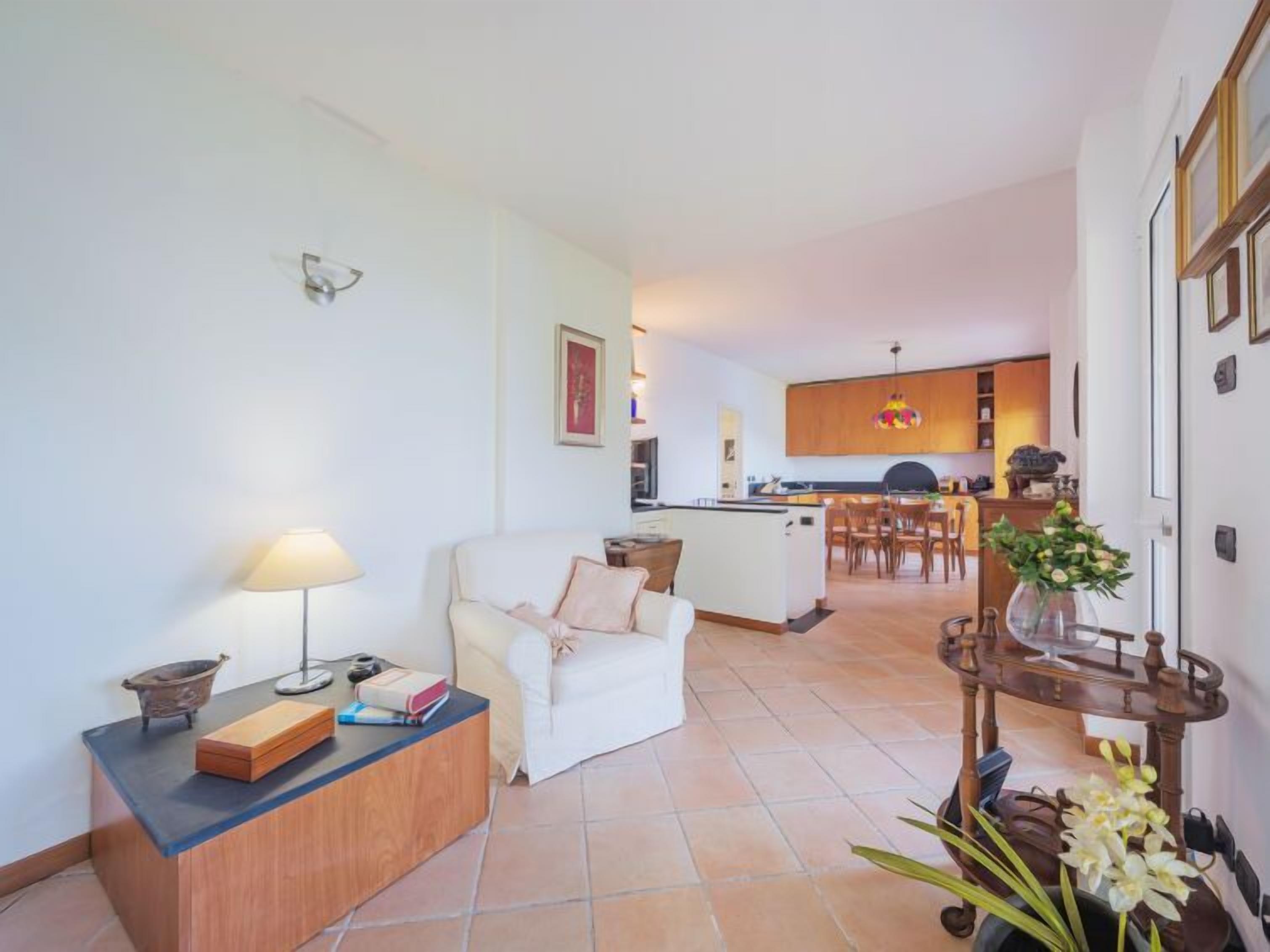 A living room with a sofa, a side table, and a lamp. There is a kitchen in the background with wooden cabinets and a dining area.