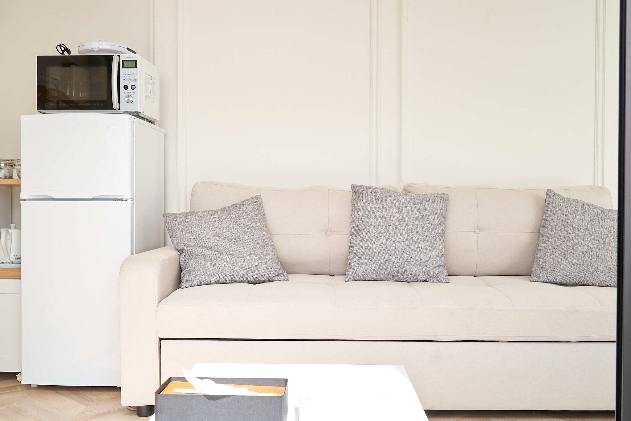 Un salon avec un canapé beige, trois coussins gris, une table basse blanche et un réfrigérateur blanc sur lequel est posé un four à micro-ondes.