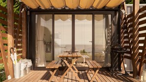 Una terraza de madera con una mesa, sillas, una parrilla y un área cubierta con vista al mar.