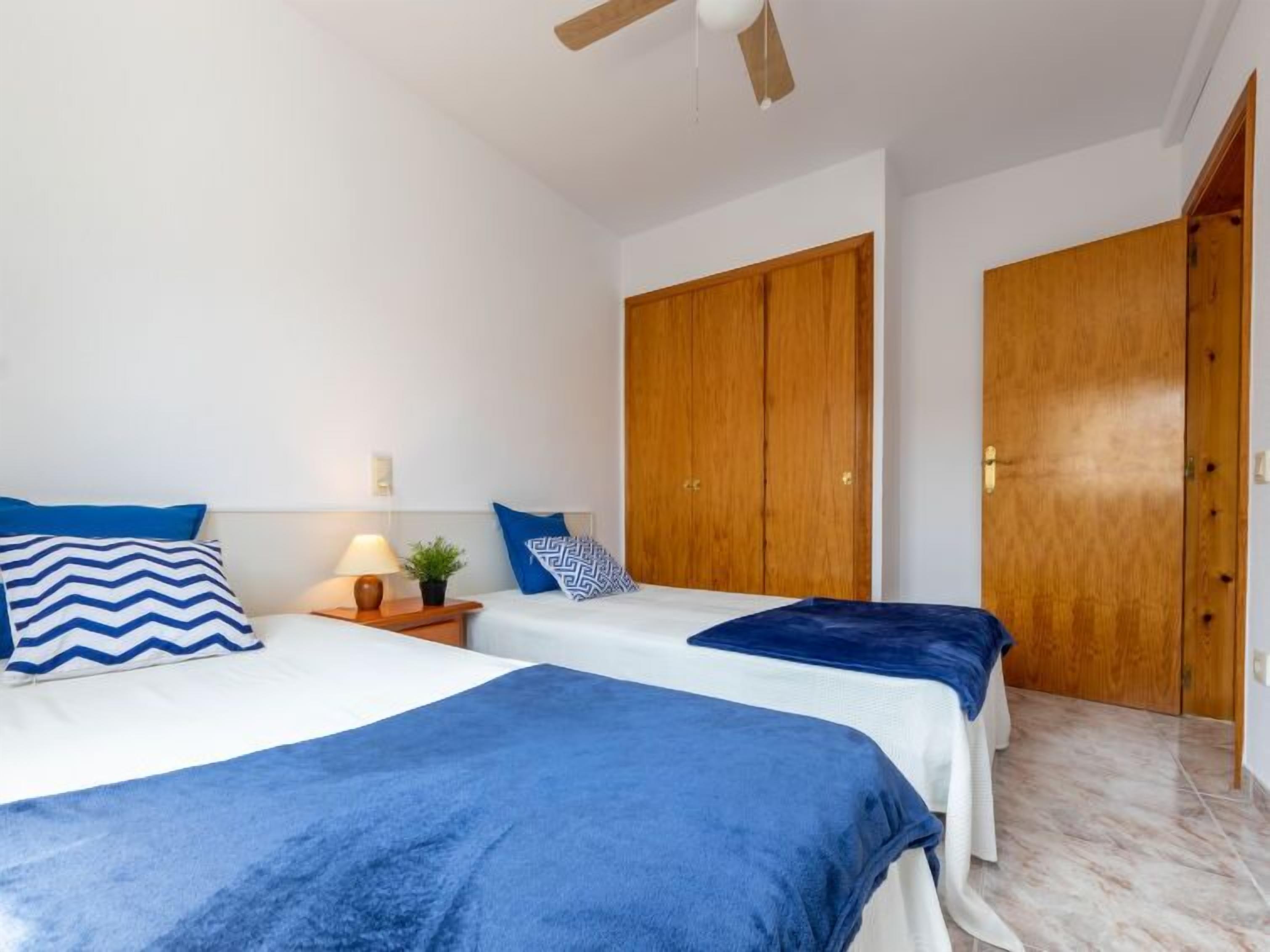 A bedroom with two beds, wooden wardrobes, a ceiling fan, and a nightstand with a lamp and a plant.